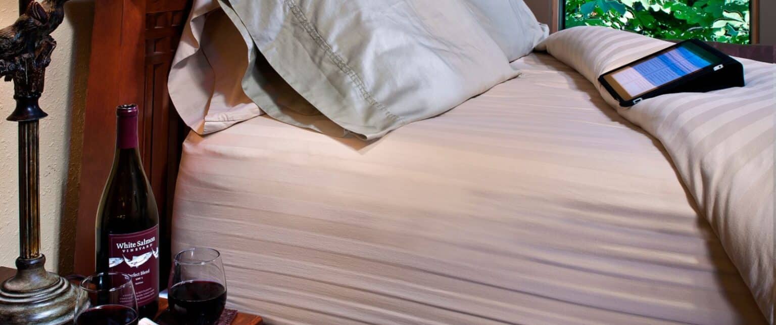 A bed with striped bedding, two wine glasses and a bottle of wine on a nightstand, and a tablet on the bed.